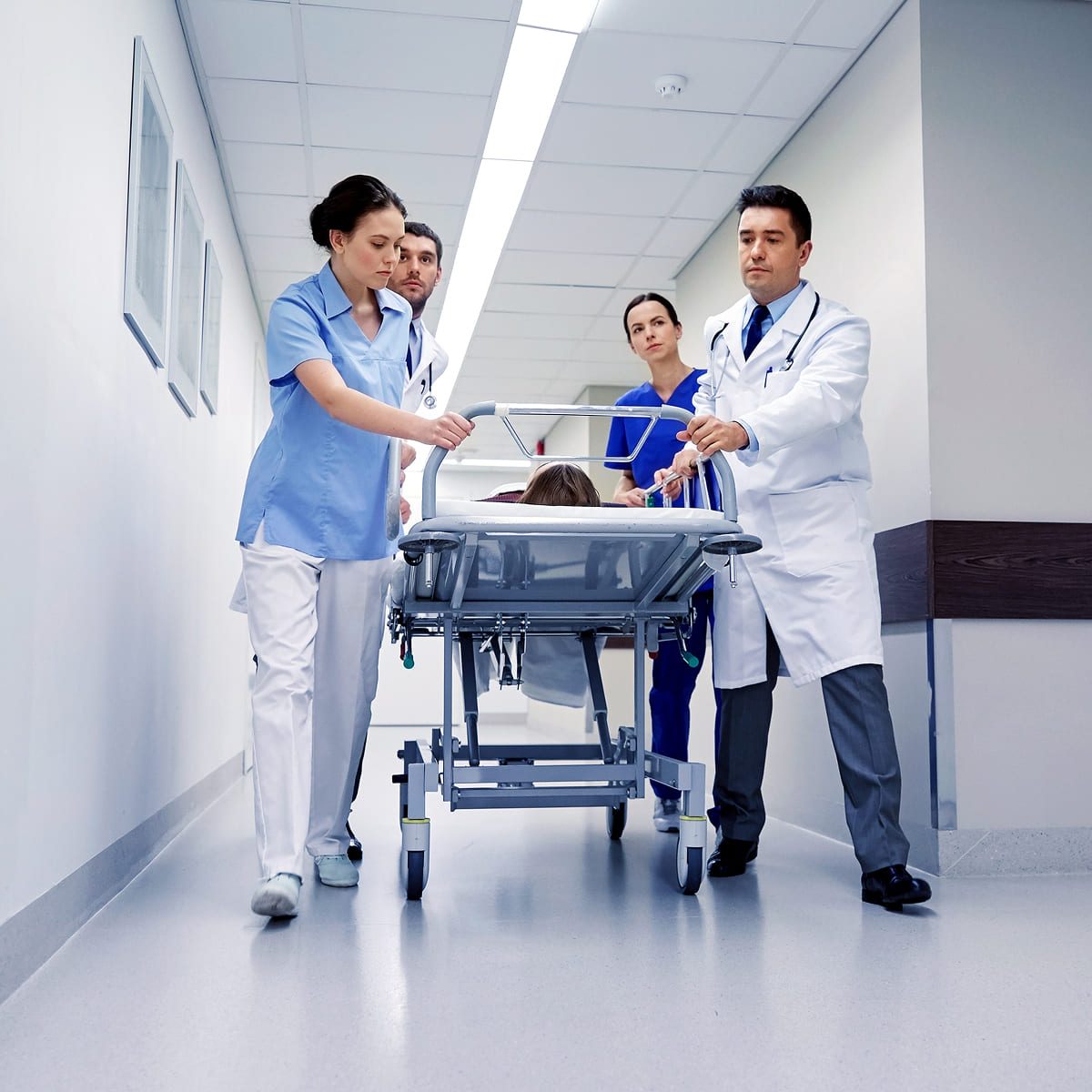 A medical team, including doctors and nurses, quickly wheels a patient on a stretcher down a hospital corridor. The atmosphere appears urgent and focused.