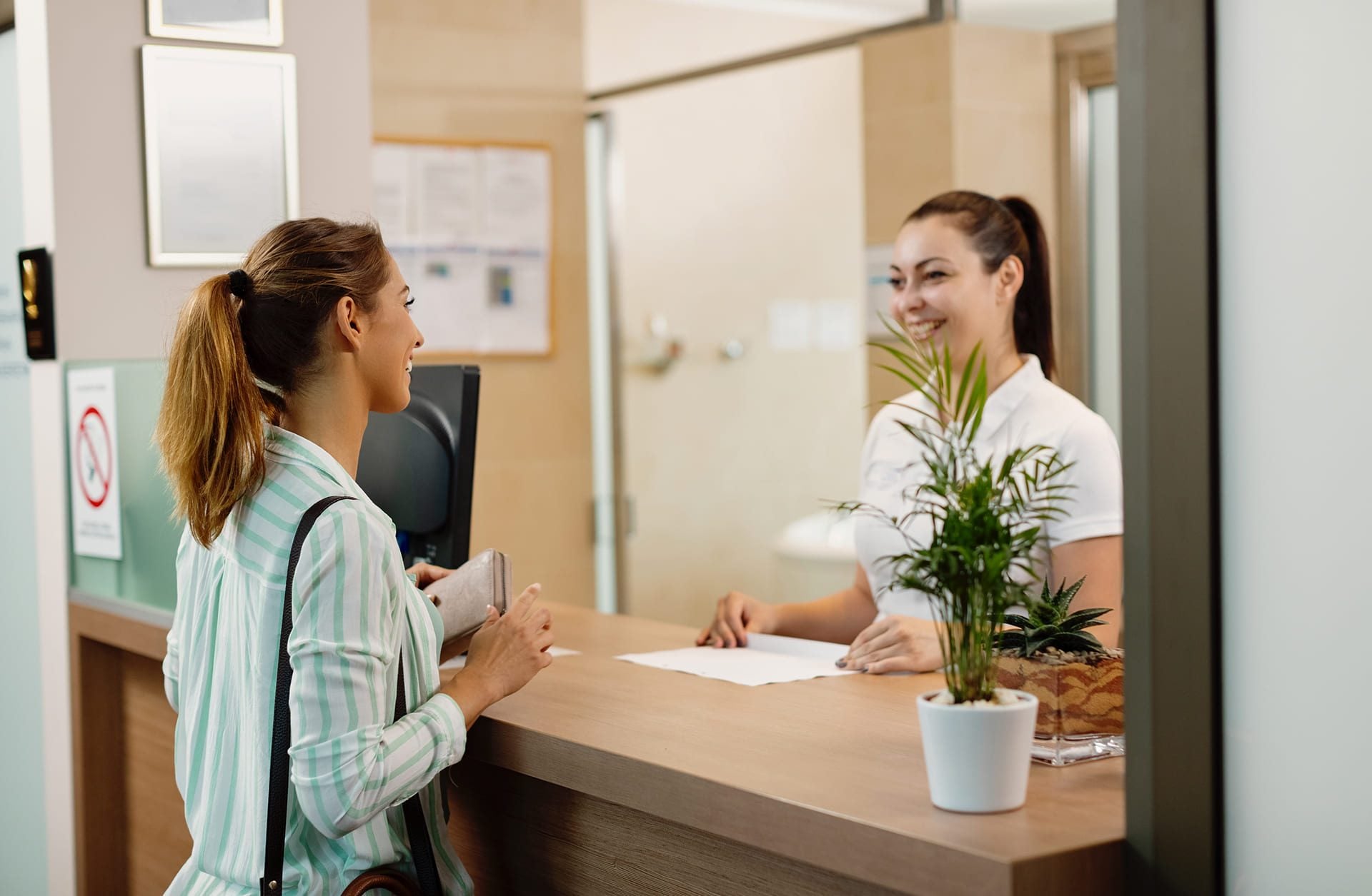 A woman stands at a reception desk holding her wallet, speaking to a smiling receptionist. There is a potted plant on the counter and documents on the wall in the background.