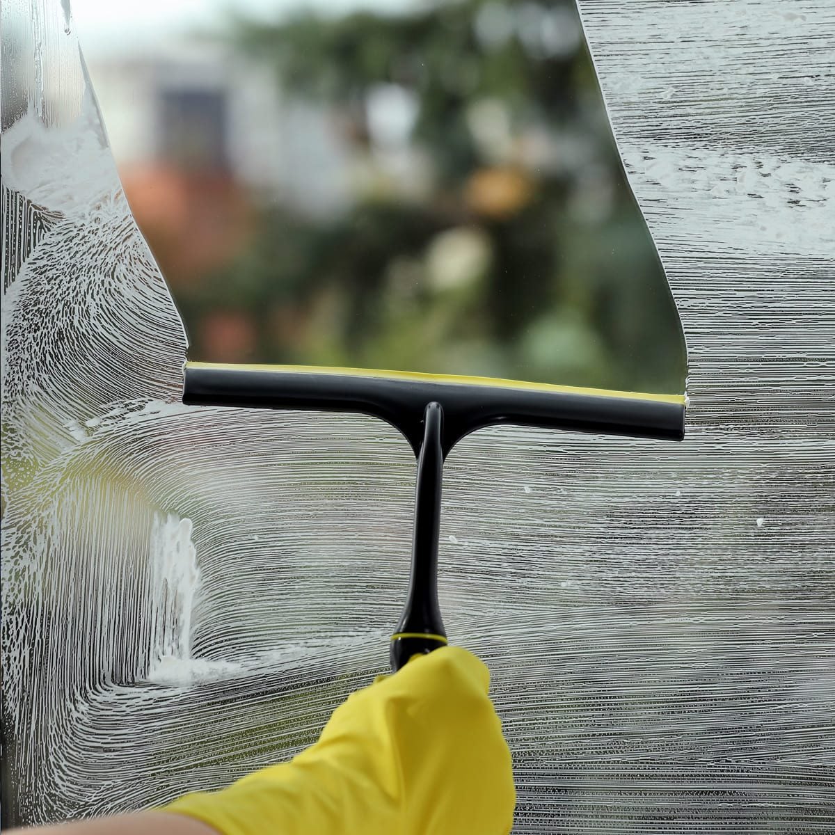 A hand wearing a yellow glove uses a squeegee to clean a soapy window, clearing part of the glass and revealing a blurred outdoor scene in the background.