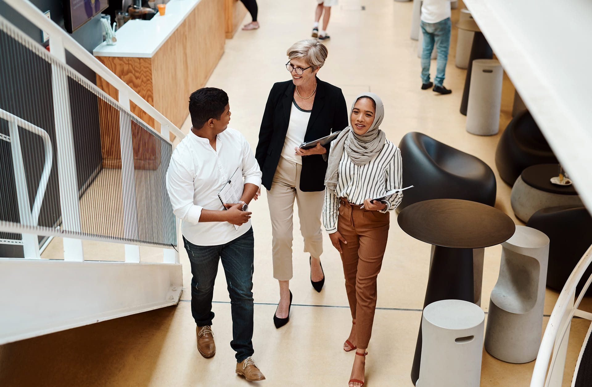 Three professionally dressed people walk indoors, smiling and talking. One holds a tablet, another a laptop. Modern furniture and a staircase are visible in the bright, open space around them.