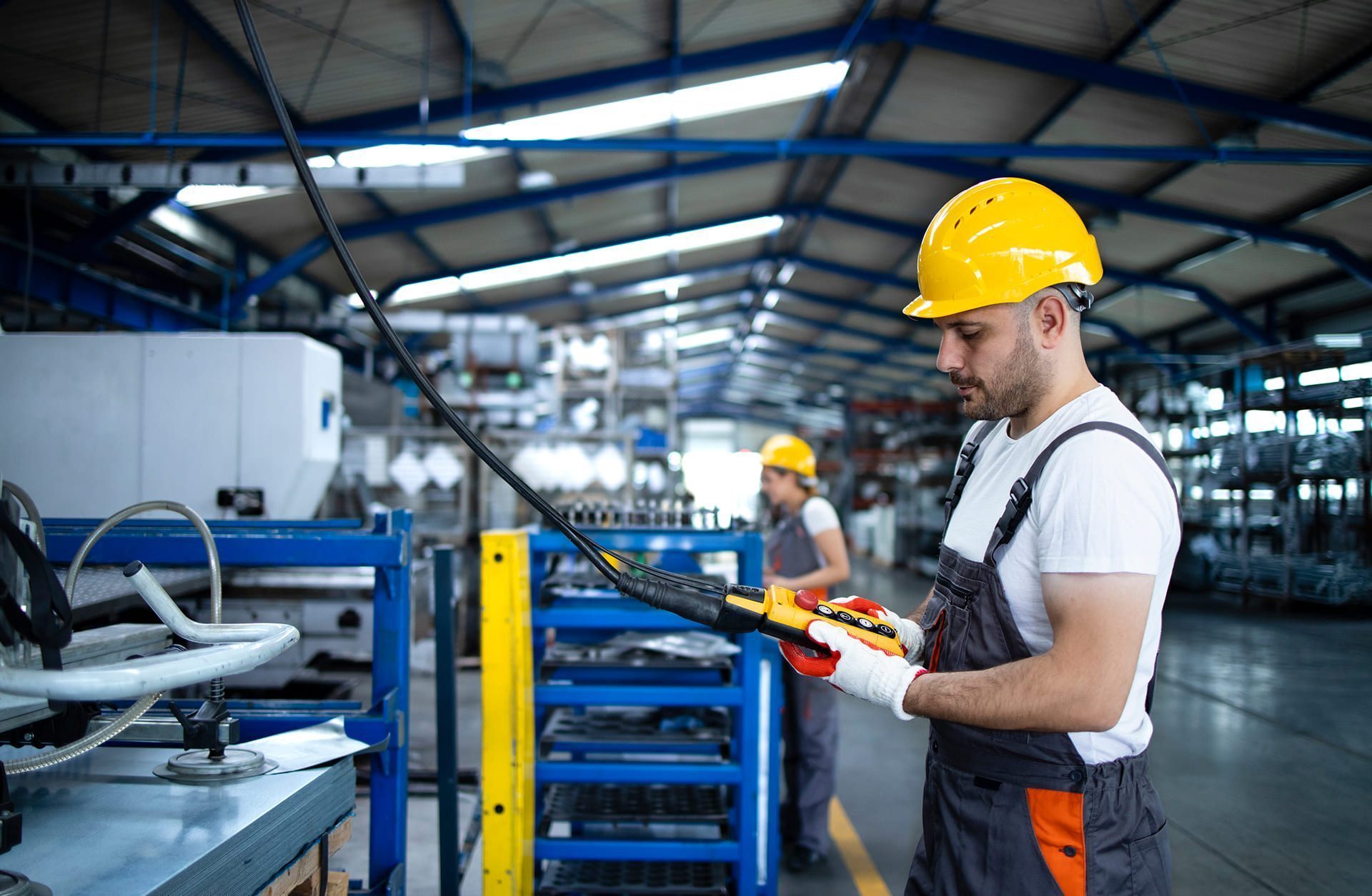 A factory worker wearing a yellow hard hat, gloves, and overalls operates machinery in an industrial setting. Shelves and equipment fill the spacious, well-lit factory, and another worker is visible in the background.