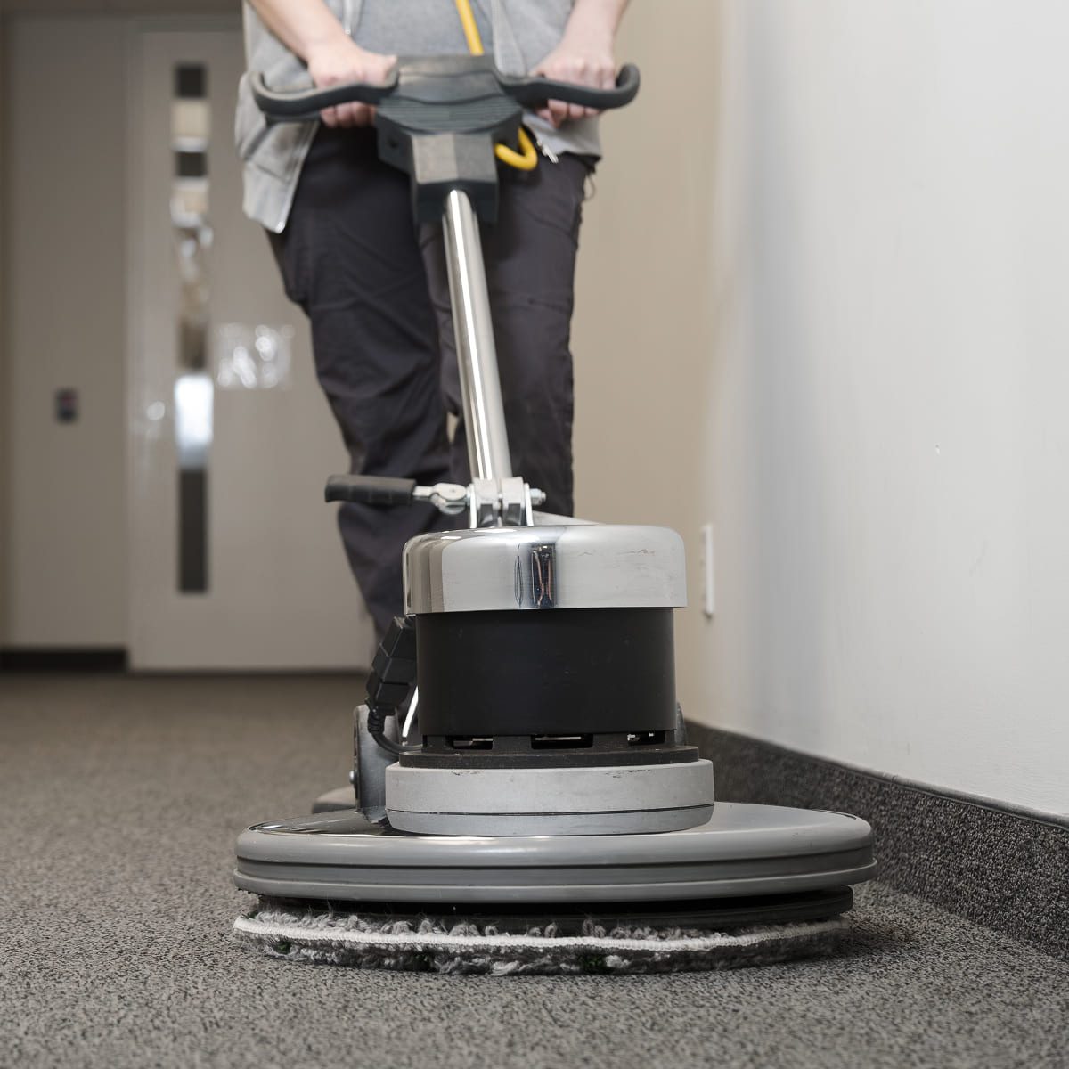 A person using a large floor cleaning machine to clean a carpeted hallway, with only their hands and lower body visible.