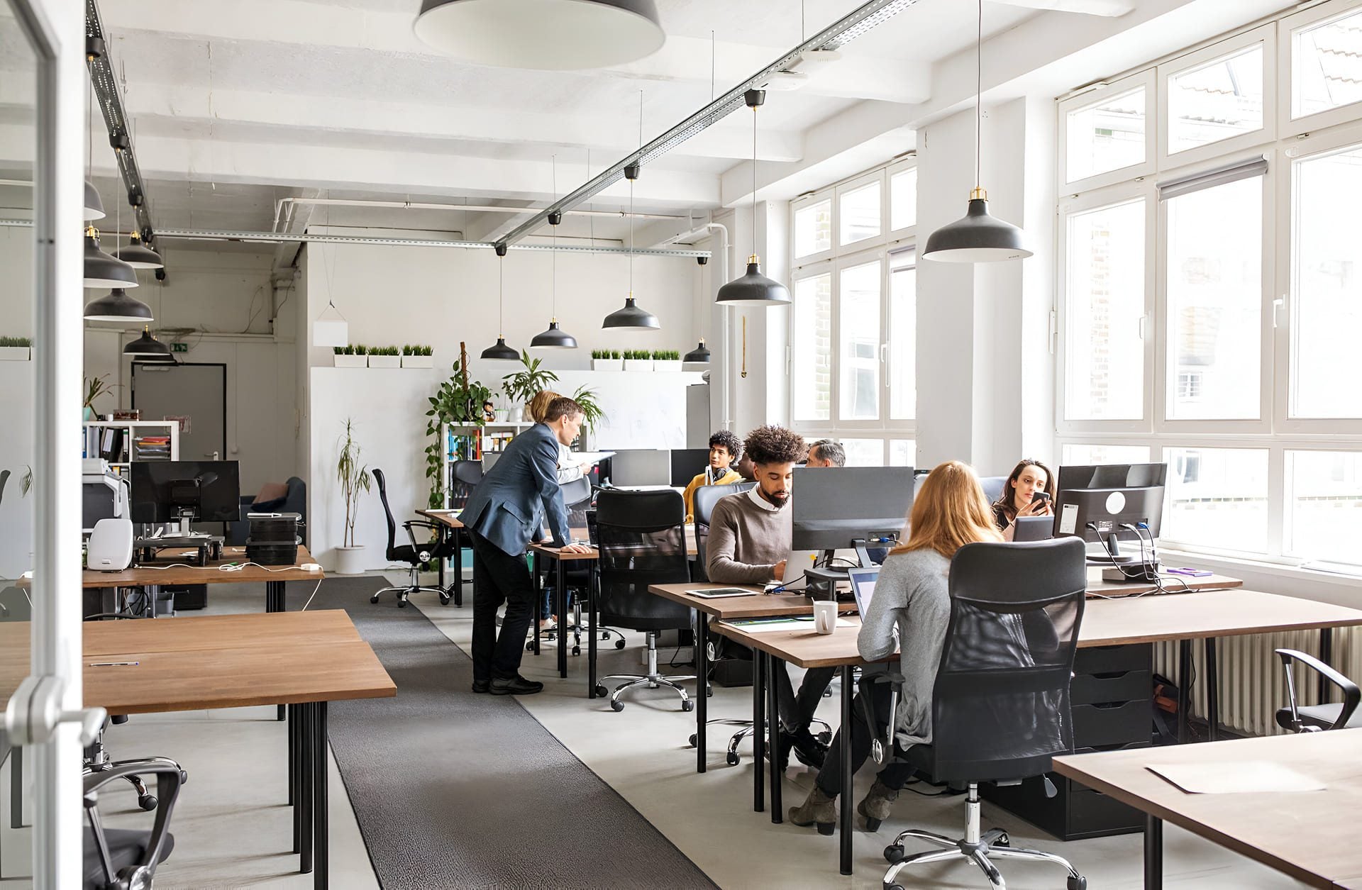 Modern open-plan office with large windows, several people working at desks with computers, bright lighting, and potted plants. One person is standing and leaning over to talk to a colleague.